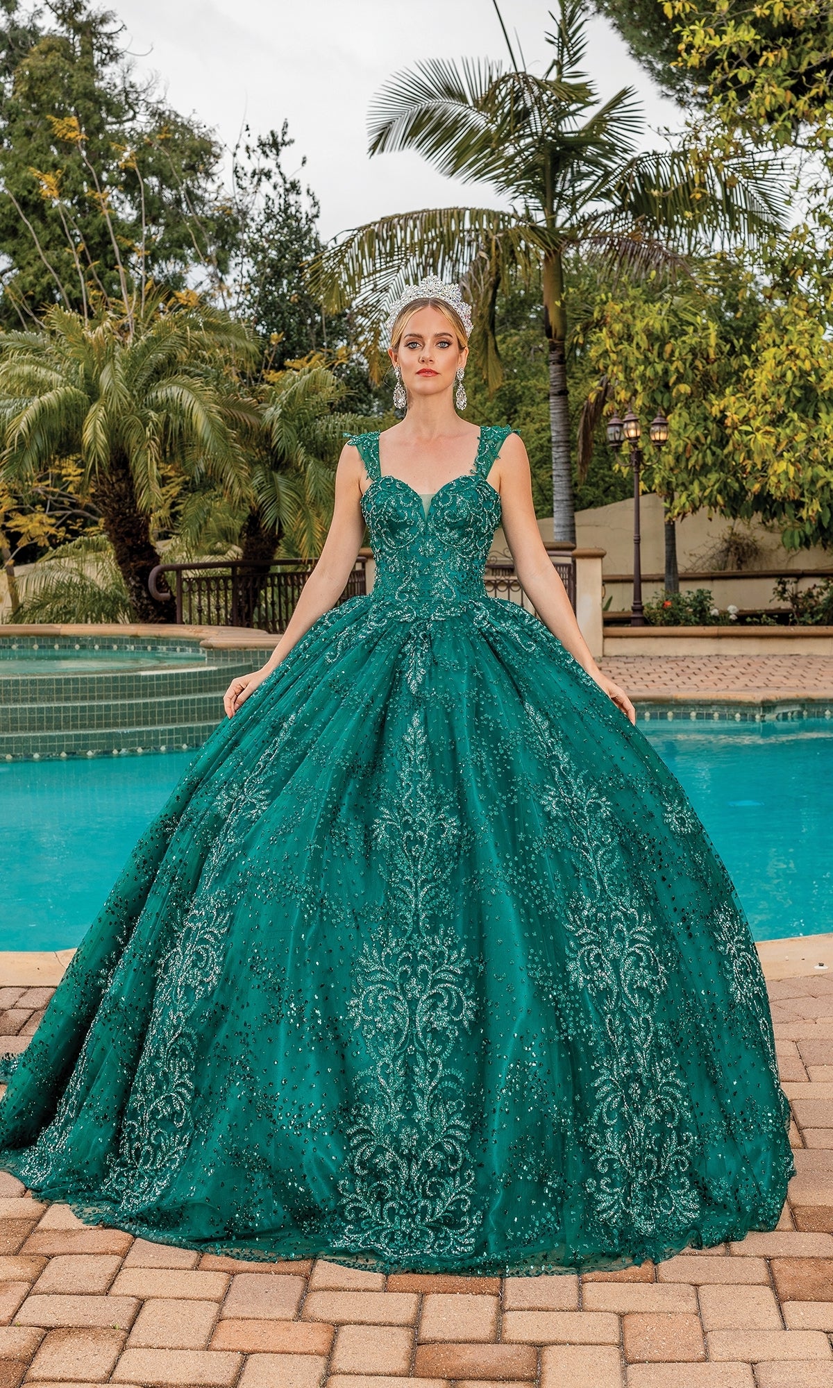 A woman stands by a pool, surrounded by palm trees and greenery, wearing the Quinceanera Dress 1784 By Dancing Queen—a teal ball gown with intricate beadwork, a sweetheart neckline, and lace embroidered details.